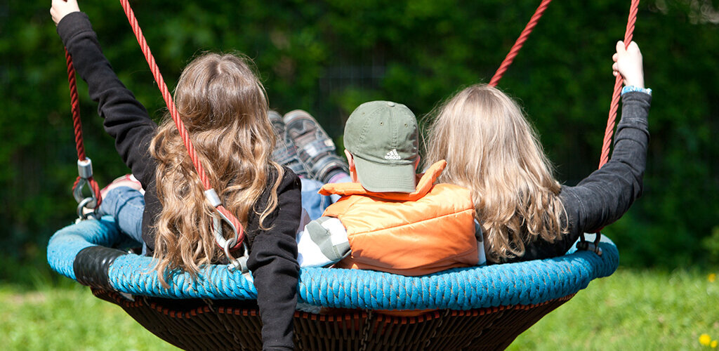 Drei Kinder sitzen mit dem Rücken zur Kamera auf einer runden Netzschaukel